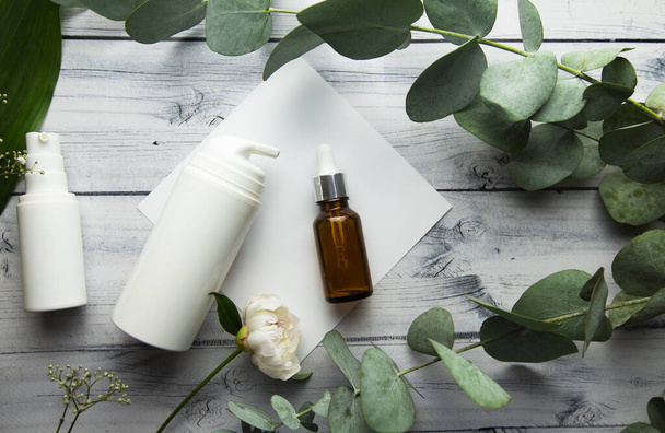 a glass bottle with hyaluronic serum and cosmetic bottles lie on a light wooden background surrounded by eucalyptus branches and plants. cosmetic product - Фото, изображение