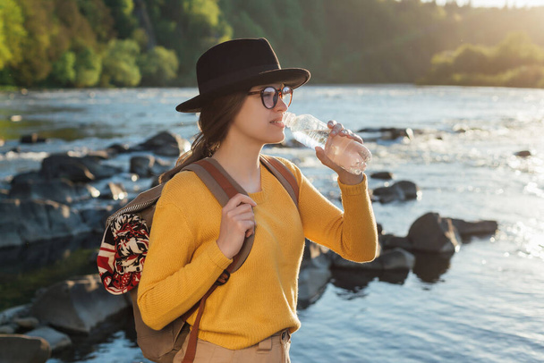 Young woman traveler drinking pure water from bottle on nature river background - Fotoğraf, Görsel