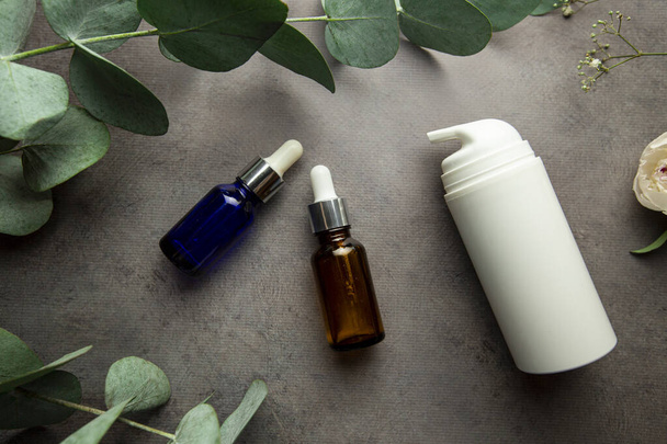flessen met serum en potjes cosmetische crèmes op een grijze achtergrond met eucalyptus takjes en groene bladeren. selectieve focus - Foto, afbeelding