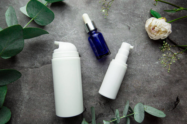 frascos con suero y frascos de cremas cosméticas sobre fondo gris con ramitas de eucalipto y hojas verdes. enfoque selectivo - Foto, Imagen