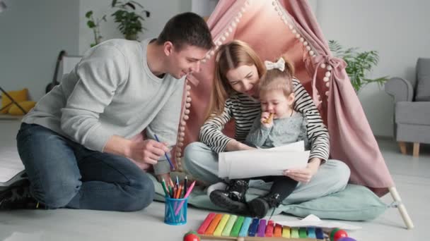 crianza de los hijos, una familia feliz con una hija pequeña viendo dibujos de niños mientras está sentada en un bigwam en casa - Metraje, vídeo