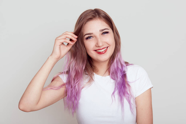 Young beautiful girl with violet hair wearing white casual T-shirt looking at camera with toothy smile and touching her hair, expressing happiness, isolated over grey background. - Фото, изображение