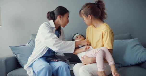 Nuori Aasia naispuolinen lastenlääkäri pidä stetoskooppi tentti pieni tyttö potilas käy lääkärin äidin kanssa istua sohvalla olohuoneessa talossa. Sairausvakuutus, Vieraile potilaan kotona käsite. - Materiaali, video