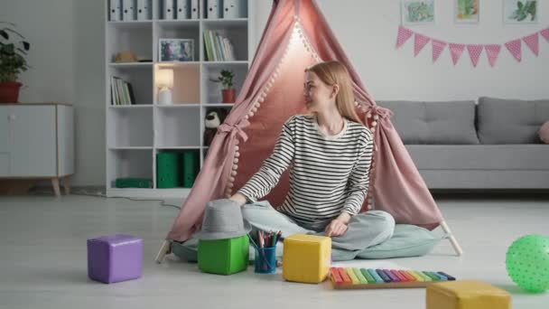 portrait of happy young female parent awaiting her beloved child while sitting in nursery background of wigwam, smiling and looking camera - Footage, Video