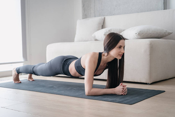 Позитивная молодая женщина с красивыми длинными волосами тренировки дома, стоя на доске предплечья на коврике йоги, чтобы тренировать мышечную силу и выносливость. Здоровый образ жизни, фитнес и тренировки - Фото, изображение