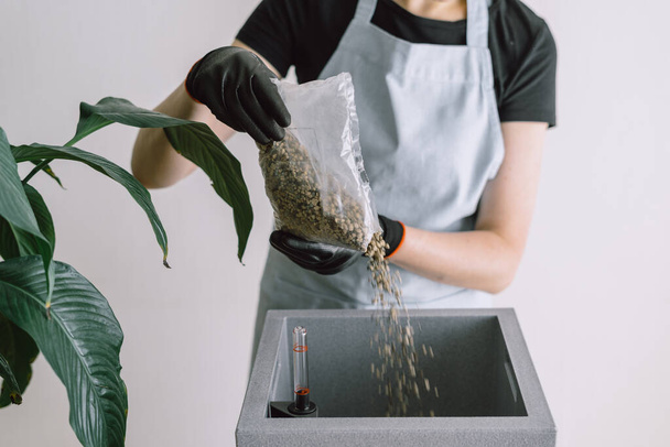 Female gardener pouring drainage with minerals to fertilize soil into prepared flower smart pot with water level indicator, standing against white background. Flowers care concept - Φωτογραφία, εικόνα