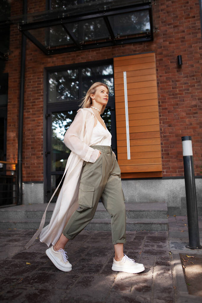 Modelo con estilo femenino Walking City Street. Fashion Woman Blogger Usando ropa de moda de primavera u otoño (gabardina beige, pantalones de carga caqui de gran tamaño, accesorios) al aire libre. Trend Street Outfit - Foto, imagen