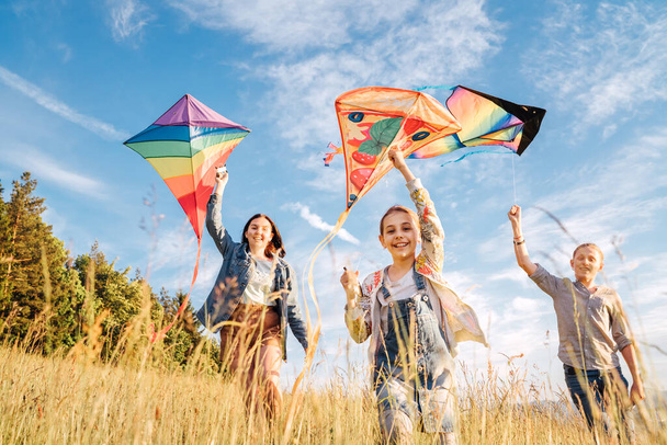 Smiling gils and brother boy running with flying colorful kites on the high grass meadow in the mountain fields. Happy childhood moments or outdoor time spending concept image.  - Fotoğraf, Görsel