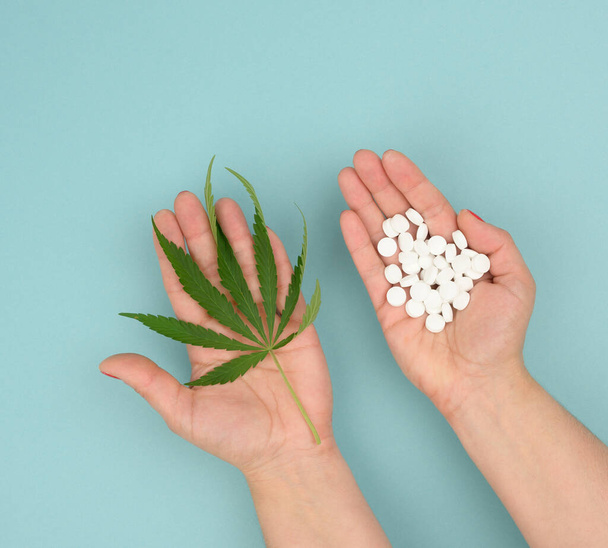 mão feminina segurando folha de cannabis verde e punhado de comprimidos redondos brancos no fundo azul, conceito de legalização como tratamento alternativo e alívio da dor - Foto, Imagem