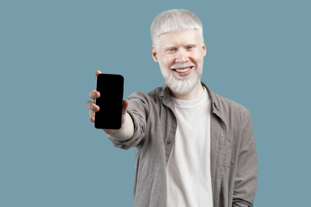 Я рекомендую це додаток. Усміхнений альбіно-хлопець тримає смартфон з порожнім екраном, ізольований на фоні бірюзової студії
 - Фото, зображення