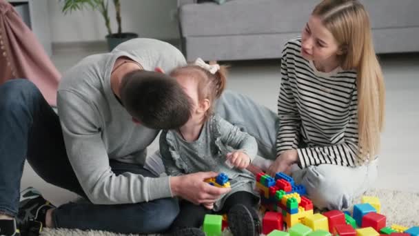 nuori rakastava perhe vanhemmat tytär nauttia yhteisestä lomasta suudella naispuolinen lapsi ja leikkii värillinen muovi rakentajat lastenhuoneessa istuu lattialla - Materiaali, video