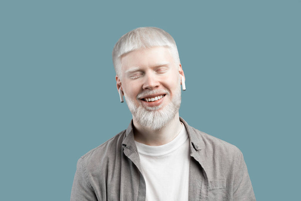 Ontspannen albino man die muziek luistert in bluetooth oordoppen, staande met gesloten ogen over turquoise achtergrond - Foto, afbeelding