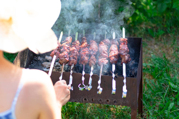 Olkihattuinen tyttö valmistaa grillillä aromaattista tuoretta lihaa vihreiden lehtien taustaa vasten kirkkaana kesäpäivänä. lähikuva - Valokuva, kuva