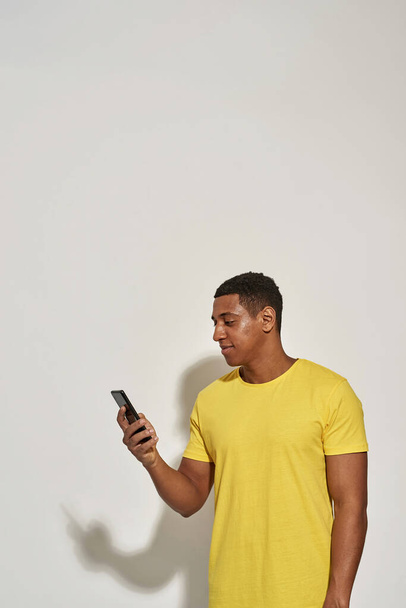 Focused young man in casual yellow t shirt looking busy, holding and using his smartphone while standing isolated over gray background - Fotó, kép