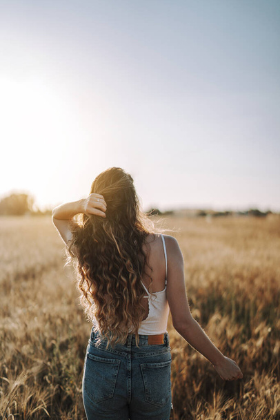 A vertical shot of a Spanish girl from behind, playing with her hair under bright sunlight in a field - Foto, imagen