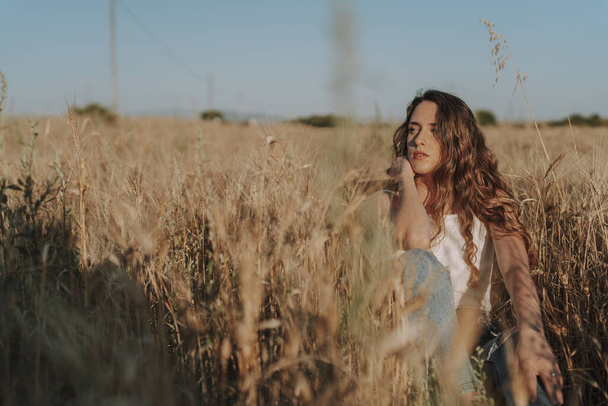 A beautiful Spanish woman sitting on the ground in a wheat field under the sun - Zdjęcie, obraz