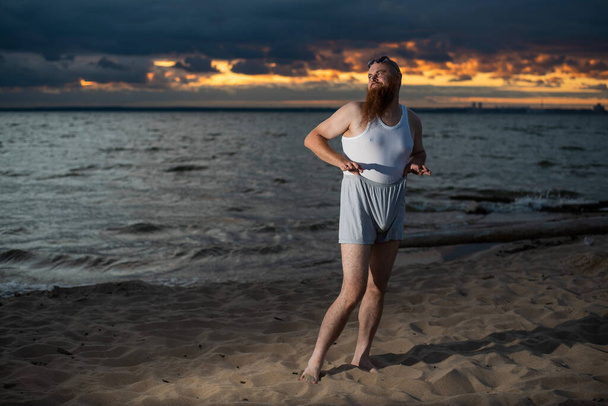 A brutal bald man posing on the beach at sunset parody glamorous chick - Fotografie, Obrázek
