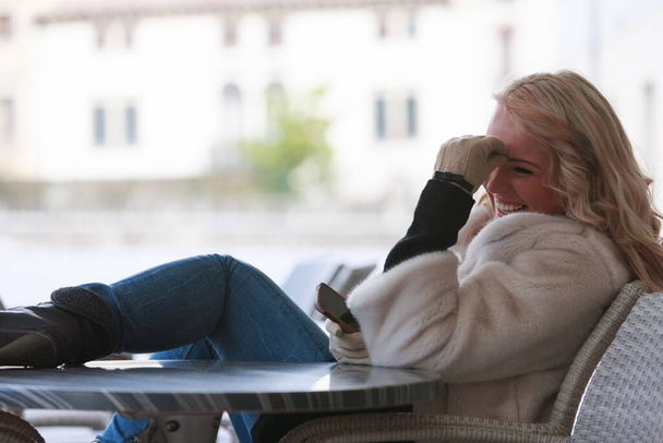 Luonnollinen muotokuva huvittunut eloisa nuori nainen nauraa iloisesti, kun hän istuu ulkona patiolla talvella matkapuhelimellaan - Valokuva, kuva