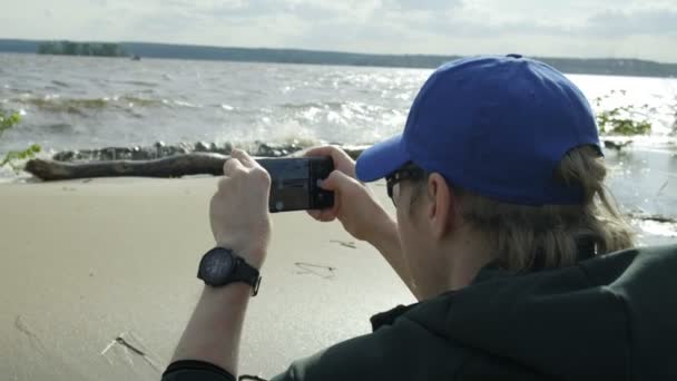 giovane uomo caucasico seduto sulla spiaggia del fiume, utilizzando lo smartphone, scattare foto - Filmati, video