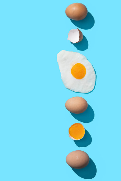 Tuoreita ruskeita munia, munankeltuaista ja paistettua munaa sinisellä pohjalla kopiointitilalla. Pystysuora muoto. - Valokuva, kuva