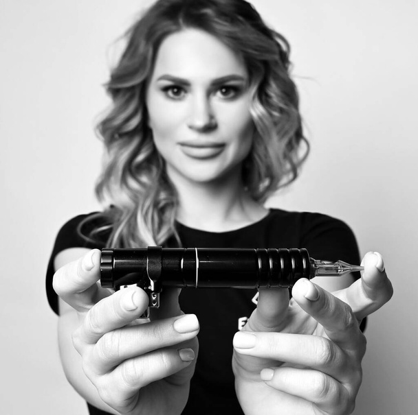 Black and white. Young curly blonde woman in black t-shirt demonstrates tattooing, permanent makeup machine, equipment - Zdjęcie, obraz