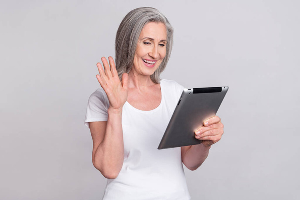 Photo of elderly woman happy positive smile talk wave hand hello video call tablet isolated over grey color background - Foto, afbeelding