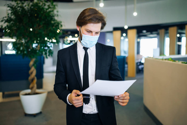 Tıbbi maskeli genç işadamı, belgelere bakıyor, modern bir ofisteki raporu kontrol ediyor.. - Fotoğraf, Görsel