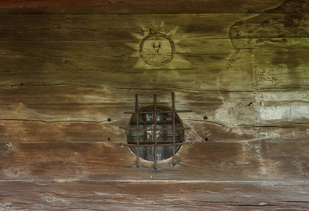 Window of round shape with metal lattice. Wooden wall with painting. Ancient orthodox wooden Ukrainian church. - Photo, Image