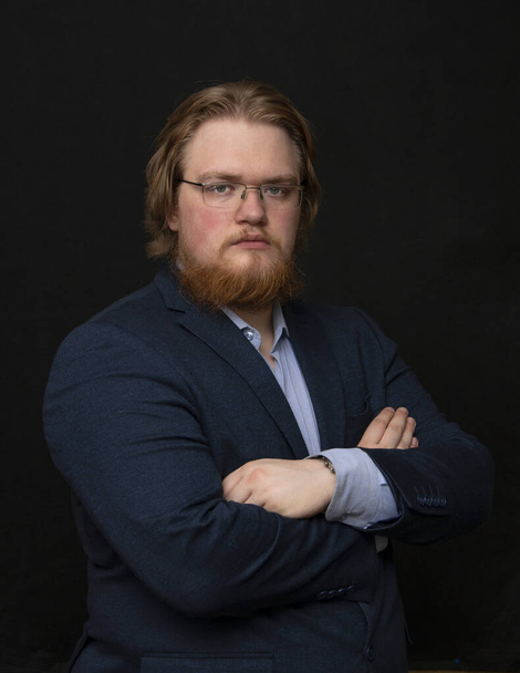 Portrait d'un homme sérieux de 25 à 30 ans avec des lunettes et un costume, avec une barbe rouge sur un fond sombre regardant l'appareil photo, en surpoids, gras, - Photo, image