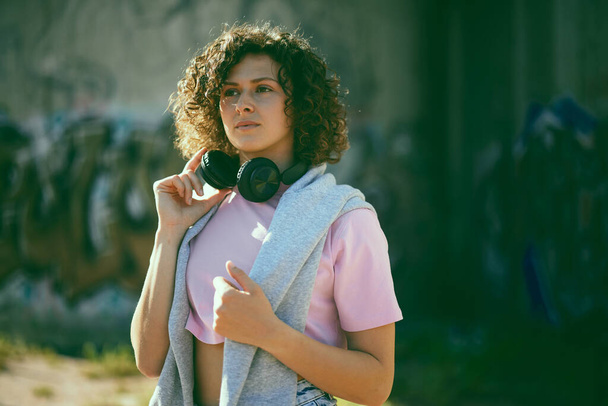 A young woman with curly hair standing outdoors and putting headphones on. - Foto, Imagen
