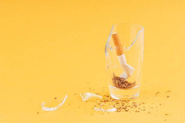 one crumpled cigarette in a broken broken glass, shards of glass are nearby. anti-smoking yellow background - Фото, зображення