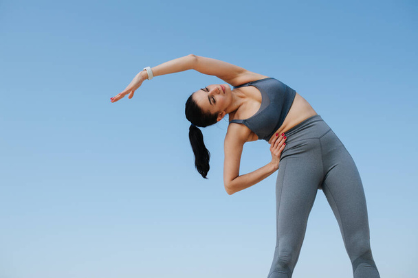 Beautiful woman in sportswear doing her stretches. Over blue sky. Upward angle - Photo, image