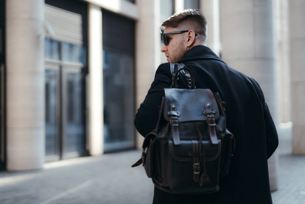 Back photo. Young businessman in a black coat and sunglasses with a backpack on his shoulder walks around the city - Photo, Image