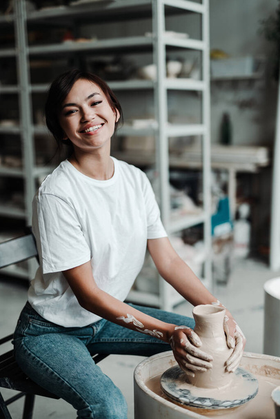Happy asian girl in a t-shirt and jeans sits on a chair makes clay dishes with her hands - Фото, изображение