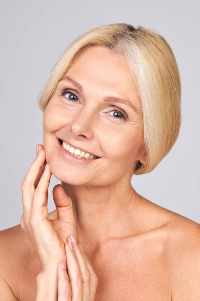 Vertical shot of smiling natural cute pretty mature Caucasian lady with blond tied hair looking at the camera and touching her cheek. Mature skin care concept for mature skin, cosmetology for mature. - Foto, afbeelding