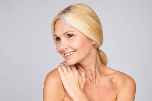 An attractive smiling natural pretty cute mature Caucasian lady, with well-groomed beautiful healthy skin, touches her shoulder with her hand and looks to the side. Mature skin care concept. - Foto, Imagen