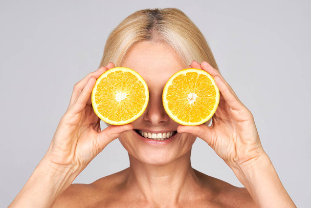 Smiling senior Caucasian blond woman with radiant skin of face holding halves of oranges like glasses, in front of her eyes, smiling cheerfully. Vitamin C cosmetics concept. Natural organic cosmetics. - Photo, Image
