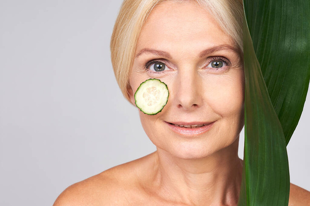 Portrait of a mature, aged beautiful woman with moisturized radiant skin, with a cucumber circle on her cheek, holding a green tropical leaf near her face and smiling tenderly. Copy space. - Fotografie, Obrázek