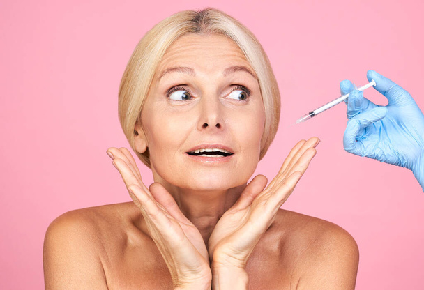 Shocked mature lady receiving beauty injections using anti-aging medication injections on pink background. Elderly woman getting collagen or hyaluronic acid injections. - Foto, Imagem