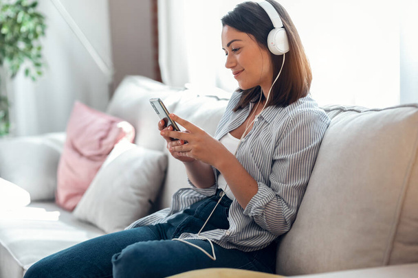 Shot of smiling young woman listening to music with smartphone while sitting on sofa at home. - Fotó, kép