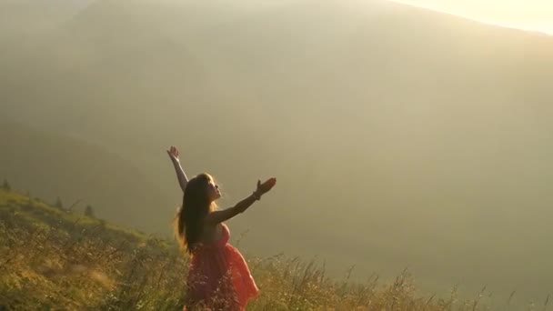 Vista posteriore di giovane donna felice viaggiatore in abito rosso in piedi su una collina erbosa in una serata ventosa in montagna d'estate con braccia tese godendo della vista della natura al tramonto. - Filmati, video