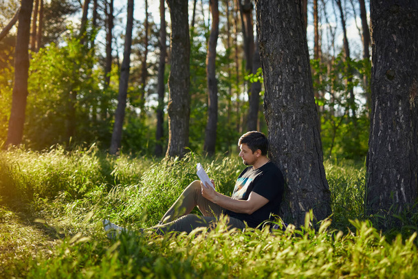 Jovem bonito sentado na floresta na grama, encostado a uma árvore, lendo um livro. Lazer exterior - Foto, Imagem