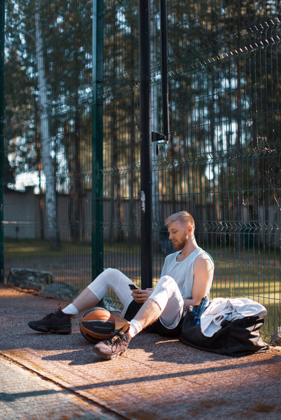 Giocatore di basket fresco giovane ragazzo avendo pausa dopo l'allenamento seduto con il telefono sul campo all'aperto in estate - Foto, immagini