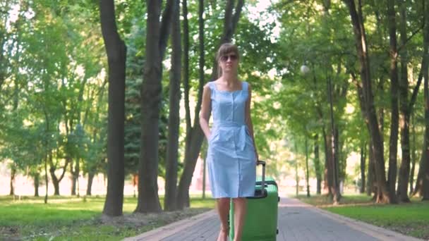 Young woman walking on park sidewalk with green suitcase on summer day. Travelling and vacation concept. - Footage, Video