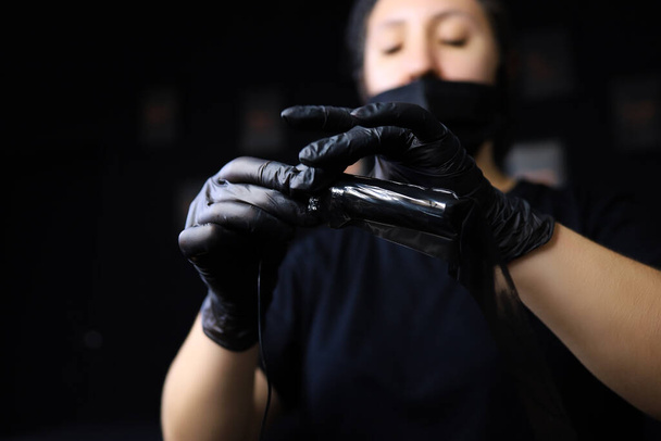 una chica que es un maestro del maquillaje permanente sostiene una máquina de tatuaje delante de ella y tira de una película protectora adicional en ella - Foto, Imagen