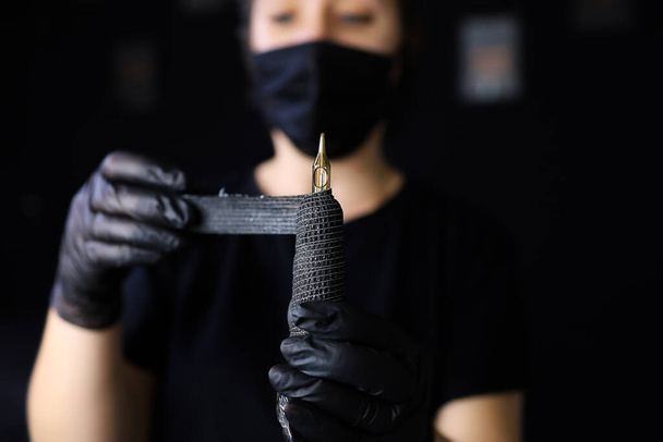 eine Tätowiererin in schwarzen Handschuhen hält ein Tätowiergerät vor sich und wickelt es zur zusätzlichen Fixierung mit einem Stretchband ein - Foto, Bild