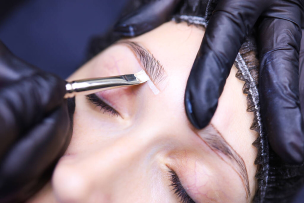the hands of the tattoo artist close up emphasize the contour of the eyebrows with a brush - Photo, Image