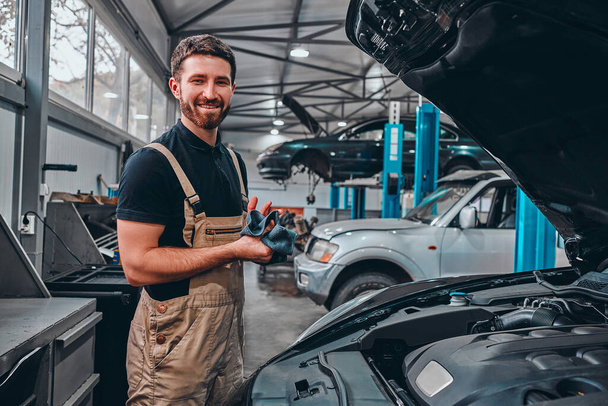 A confident young man in uniform wiping his hands with a rag and smiling while standing in the workshop with a car on the background. - Фото, зображення