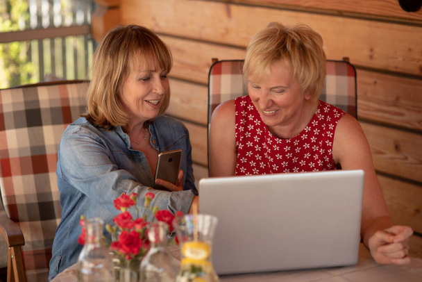 Starsze kobiety śmieją się razem korzystając z laptopa i oglądając go w domu. - Zdjęcie, obraz