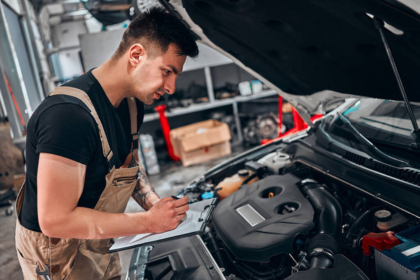 Car service inventory. Mechanic in auto repair shop makes a note on a clipboard while checking on a car engine. Side view. - Φωτογραφία, εικόνα
